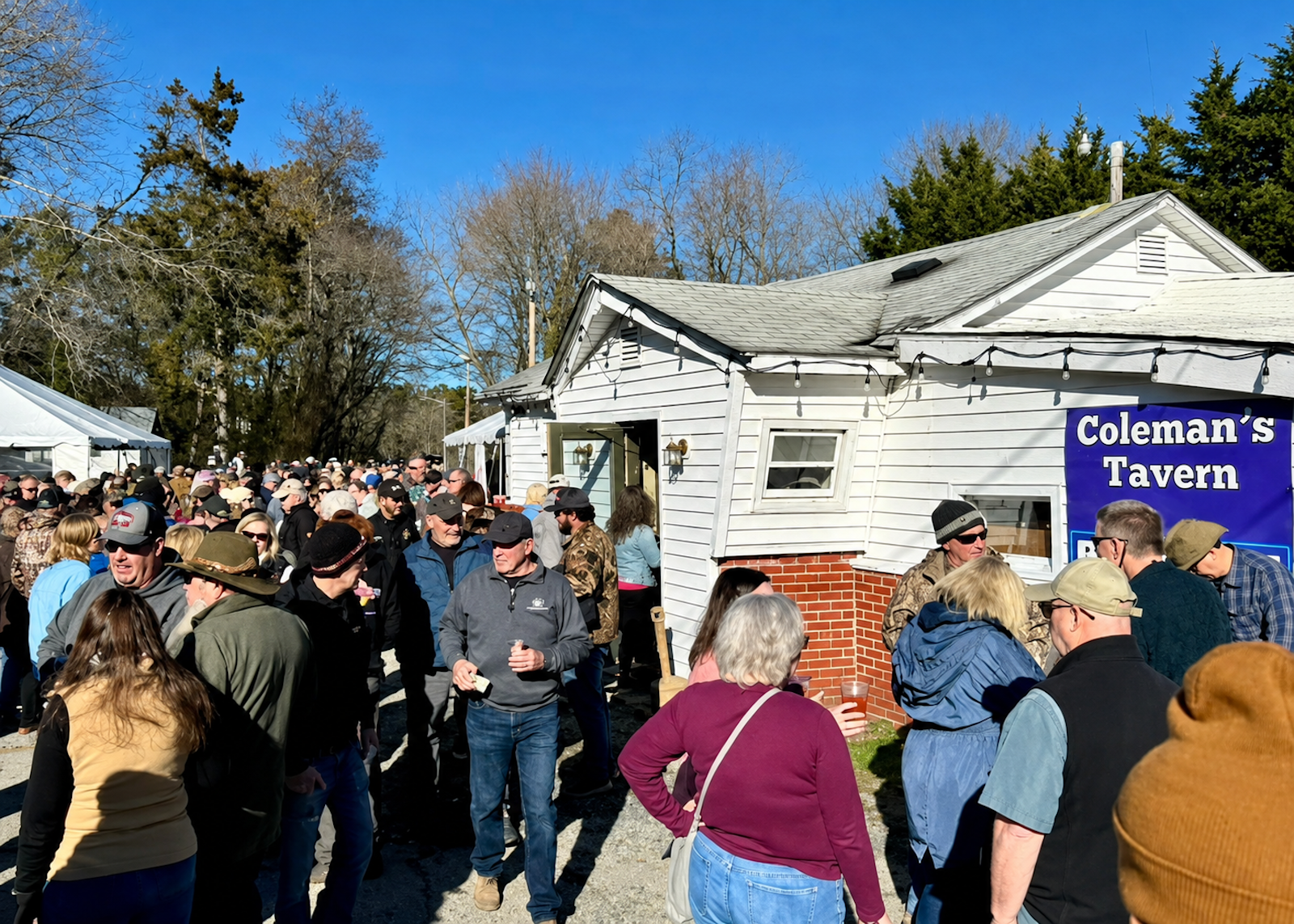 Large crowd gathered outside Coleman's Tavern, people are standing and socializing on a sunny day