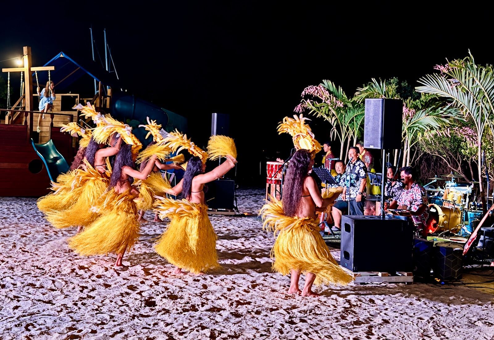 Hula dancers in yellow skirts performing on a beach at night beside a live band and palm trees.
