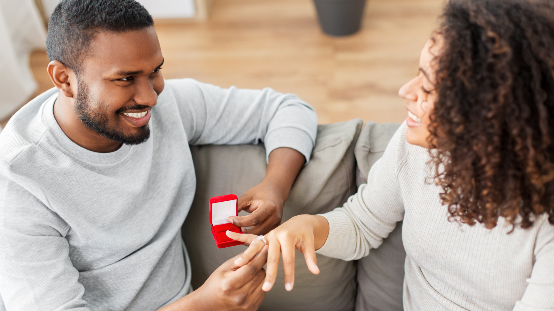 Man proposing to woman with a ring on a sofa; both smile, red box.