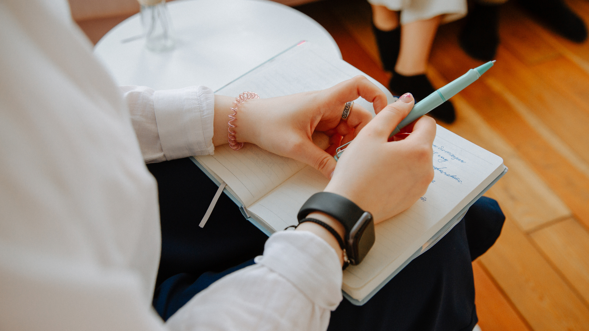 Person writing in a notebook, wearing a smartwatch and bracelet. Light blue pen in hand.