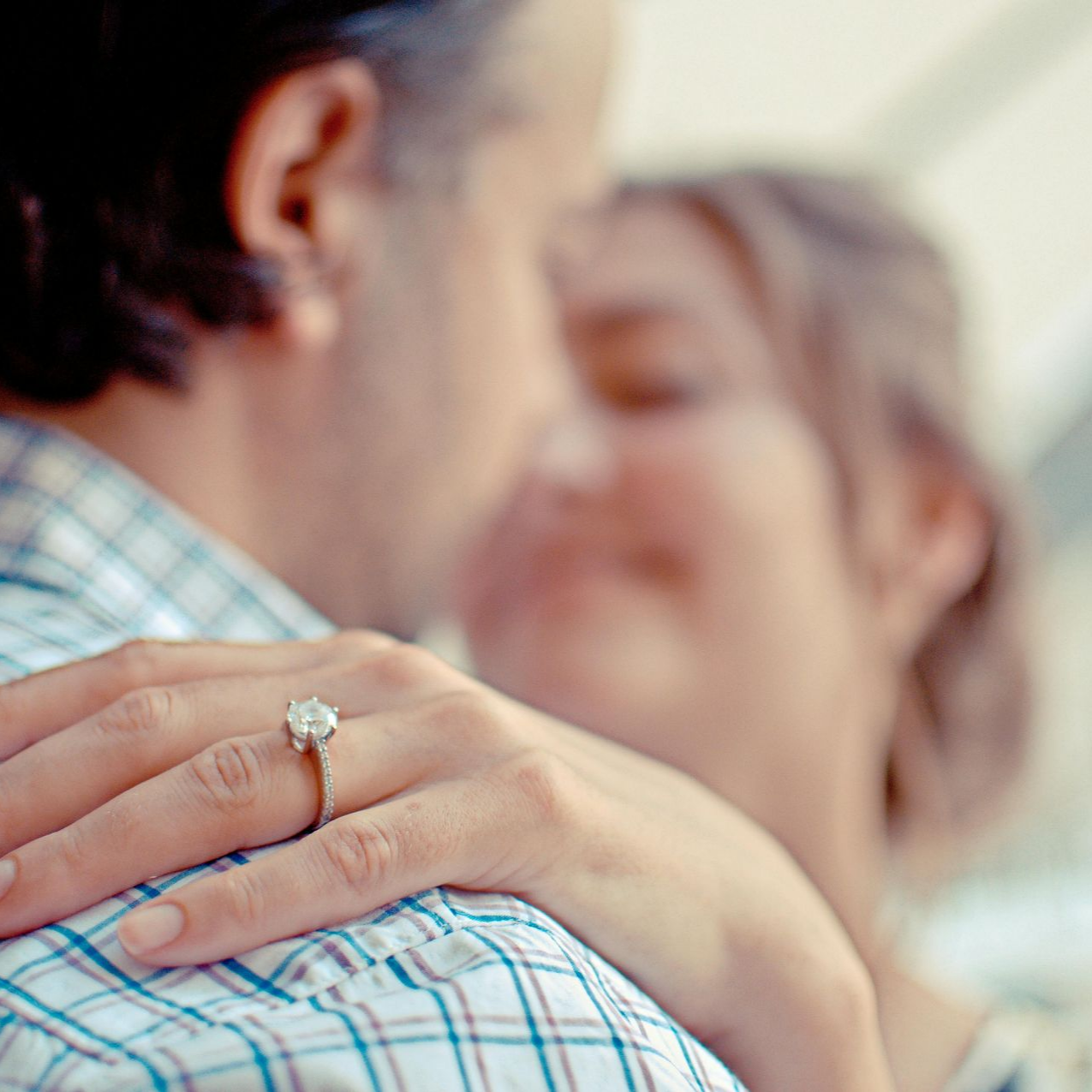 Man embraces woman, her hand with engagement ring on his shoulder.