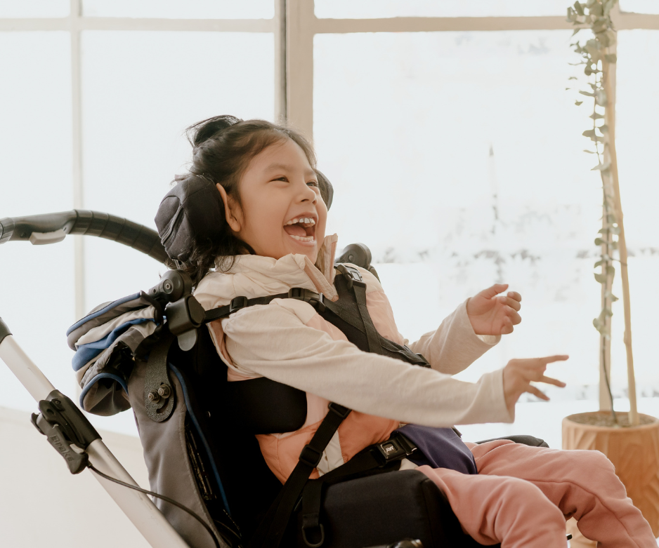 Un bambino che ride, seduto su una sedia a rotelle, si protende in avanti in una stanza luminosa vicino a una finestra.