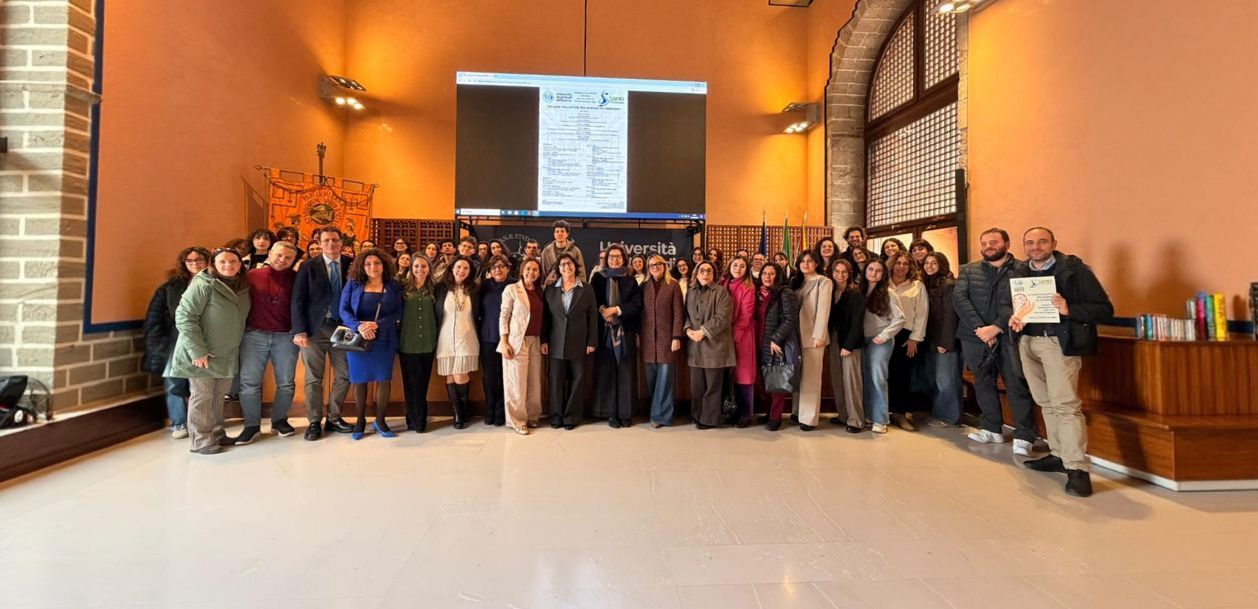 Un folto gruppo di persone posa in una sala illuminata da una luce calda, con uno schermo per proiezioni alle loro spalle.