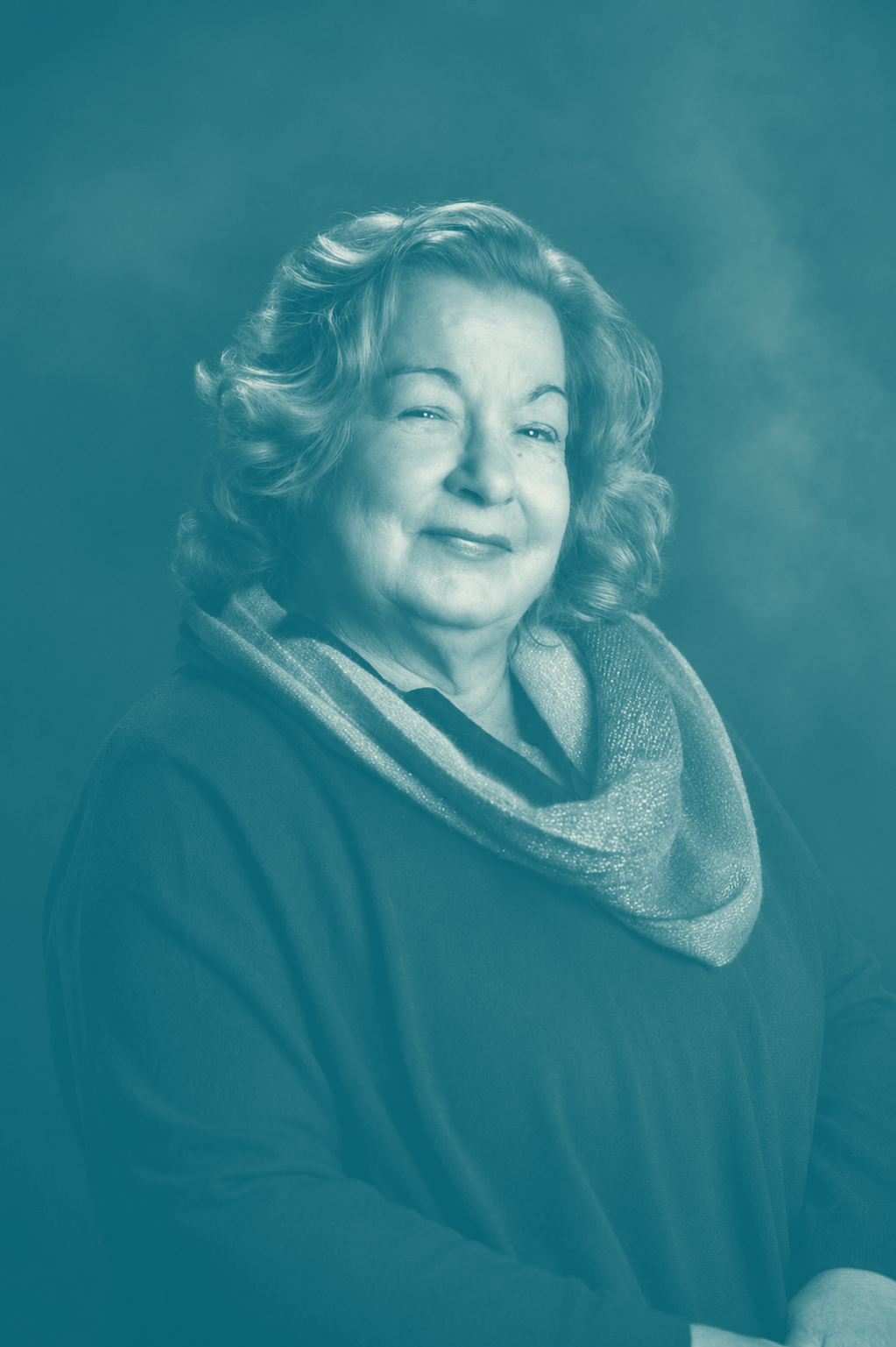 Ritratto di una persona sorridente con indosso una maglia scura e una sciarpa fantasia, su uno sfondo color verde acqua in studio.
