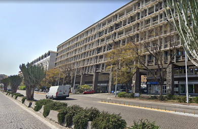 Moderno edificio per uffici che si affaccia su un'ampia strada con furgoni parcheggiati e alberi sotto un cielo limpido.
