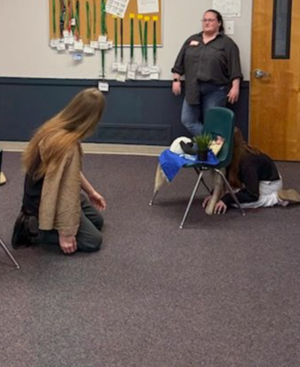 Two students kneel on carpet beside a chair and desk in a classroom while an adult stands by the door.