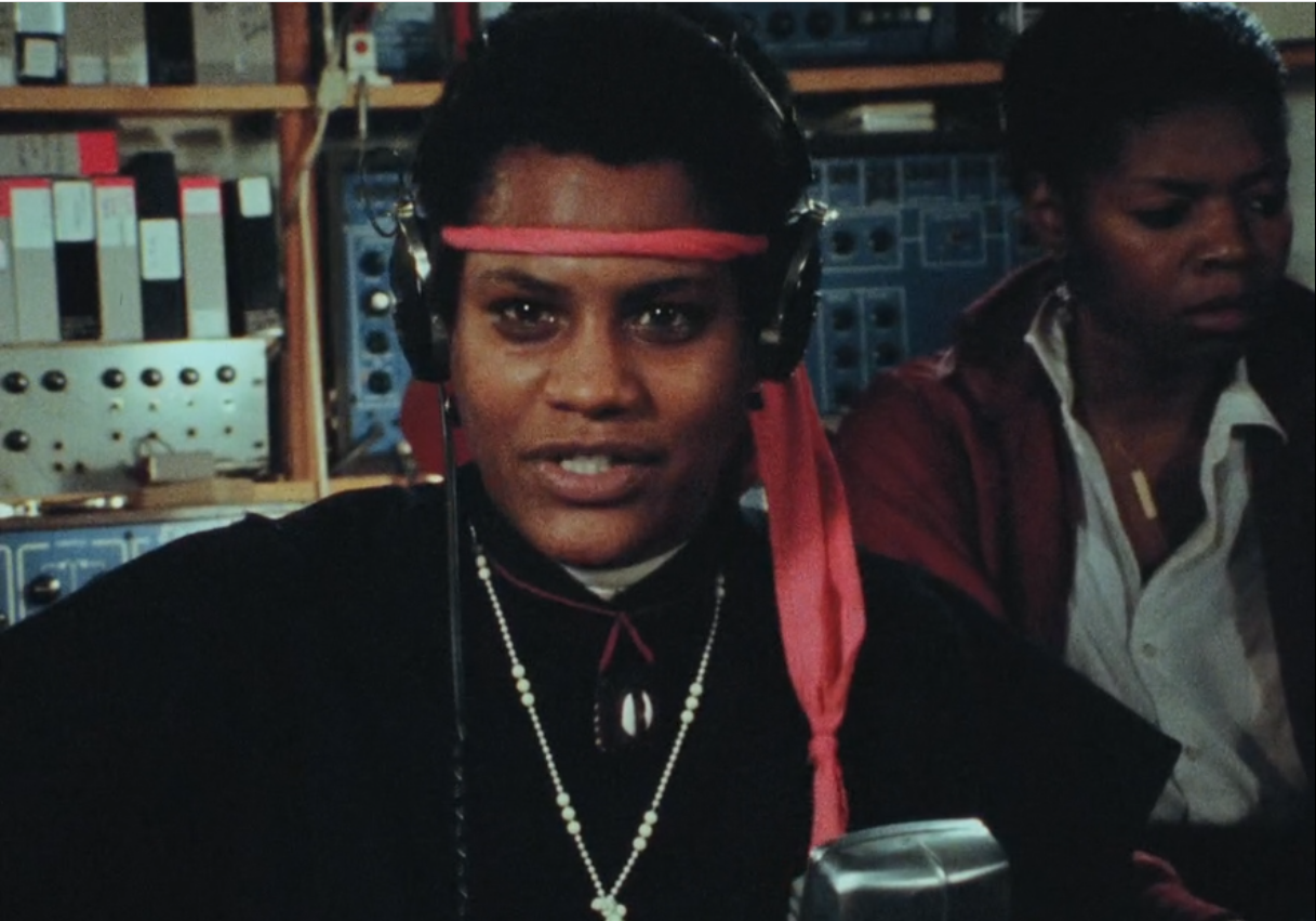 Person in a red headband and necklaces sits in front of shelves, with another person in the background.