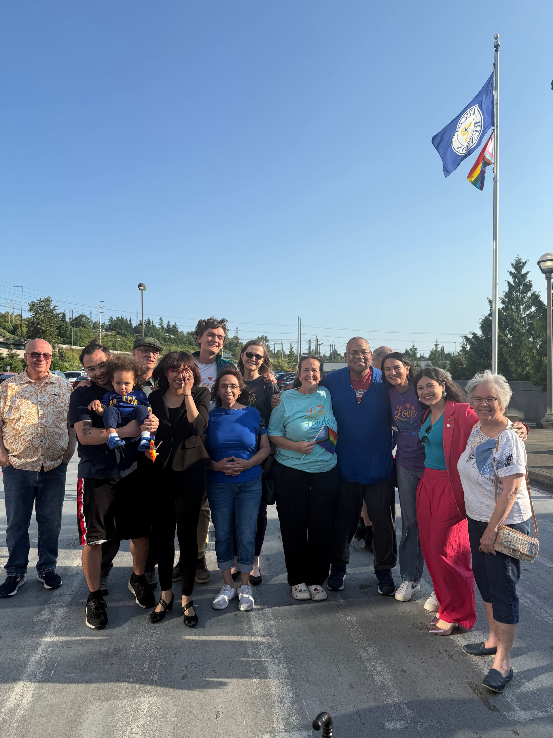 A group of people of a variety of ages, sizes, and colors are posing for a picture in front of a flag pole.