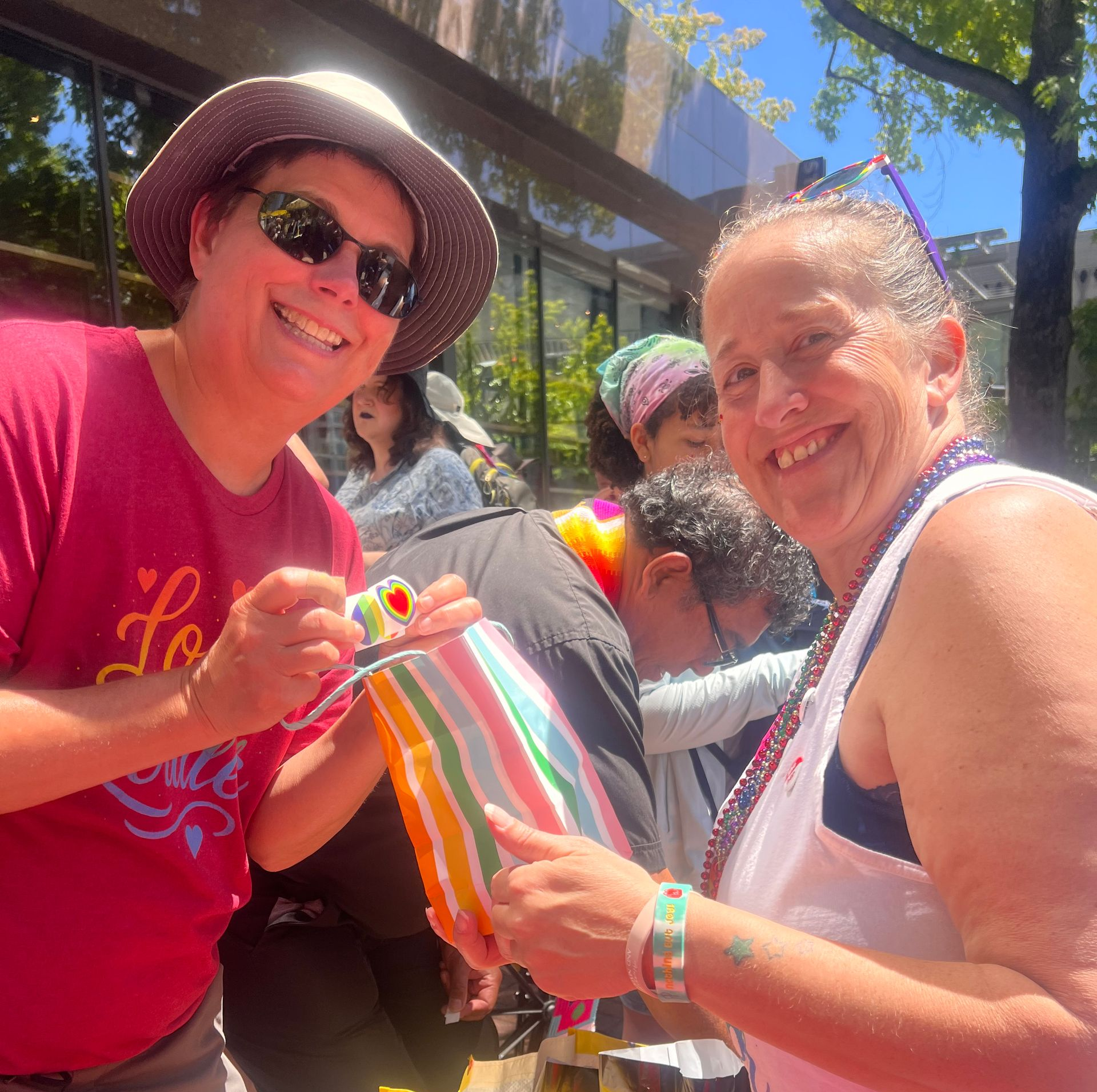 Two smiling people at an outdoor event hold a rainbow-striped bag in bright sunlight