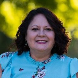 A Latina woman in a blue floral shirt is smiling for the camera.