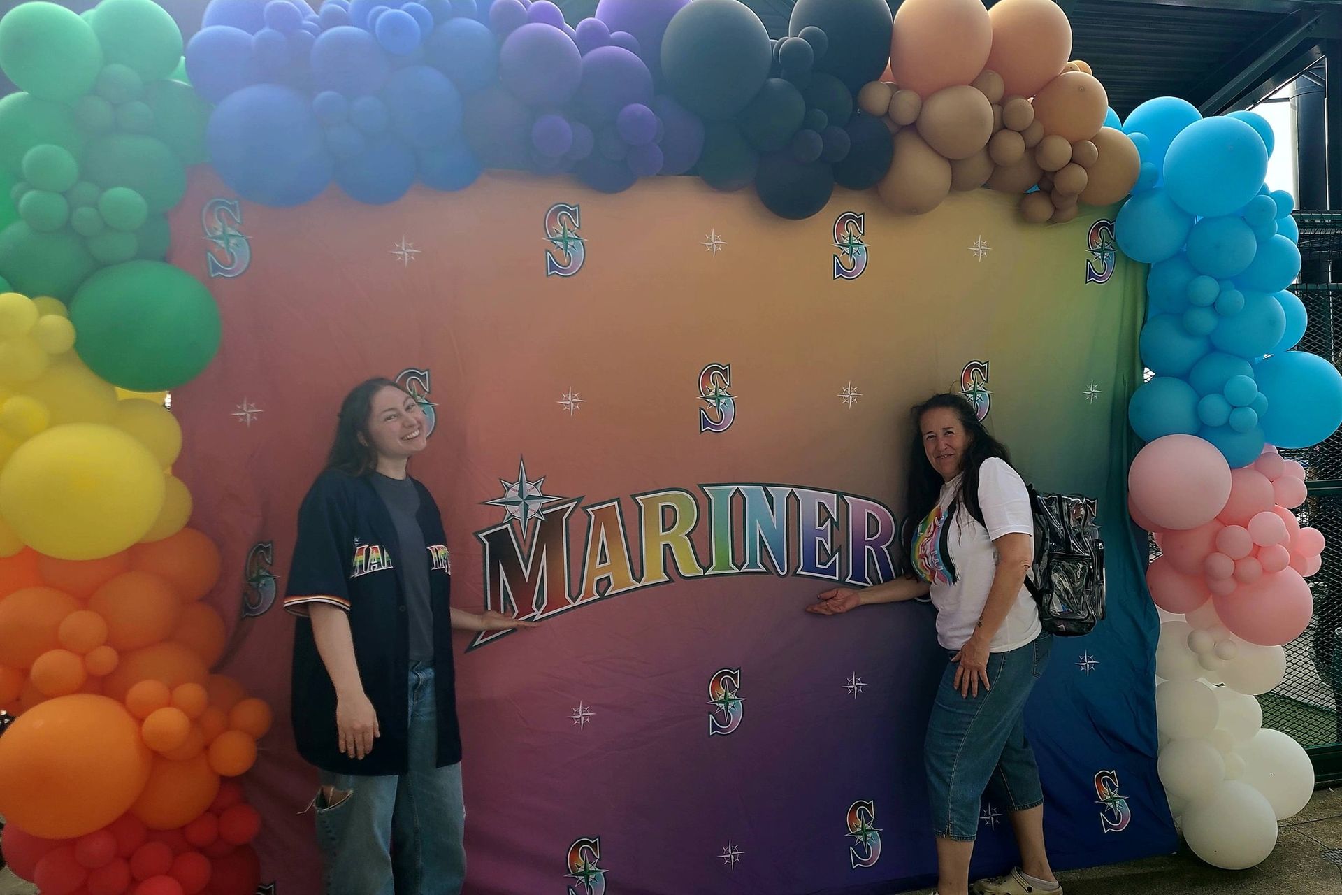 Two people posing beside a colorful balloon arch and “MARINERS” backdrop