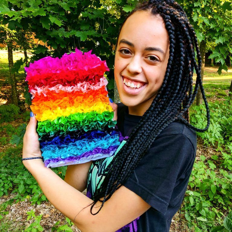 Person smiling and holding a rainbow-colored layered cake outdoors in a green park