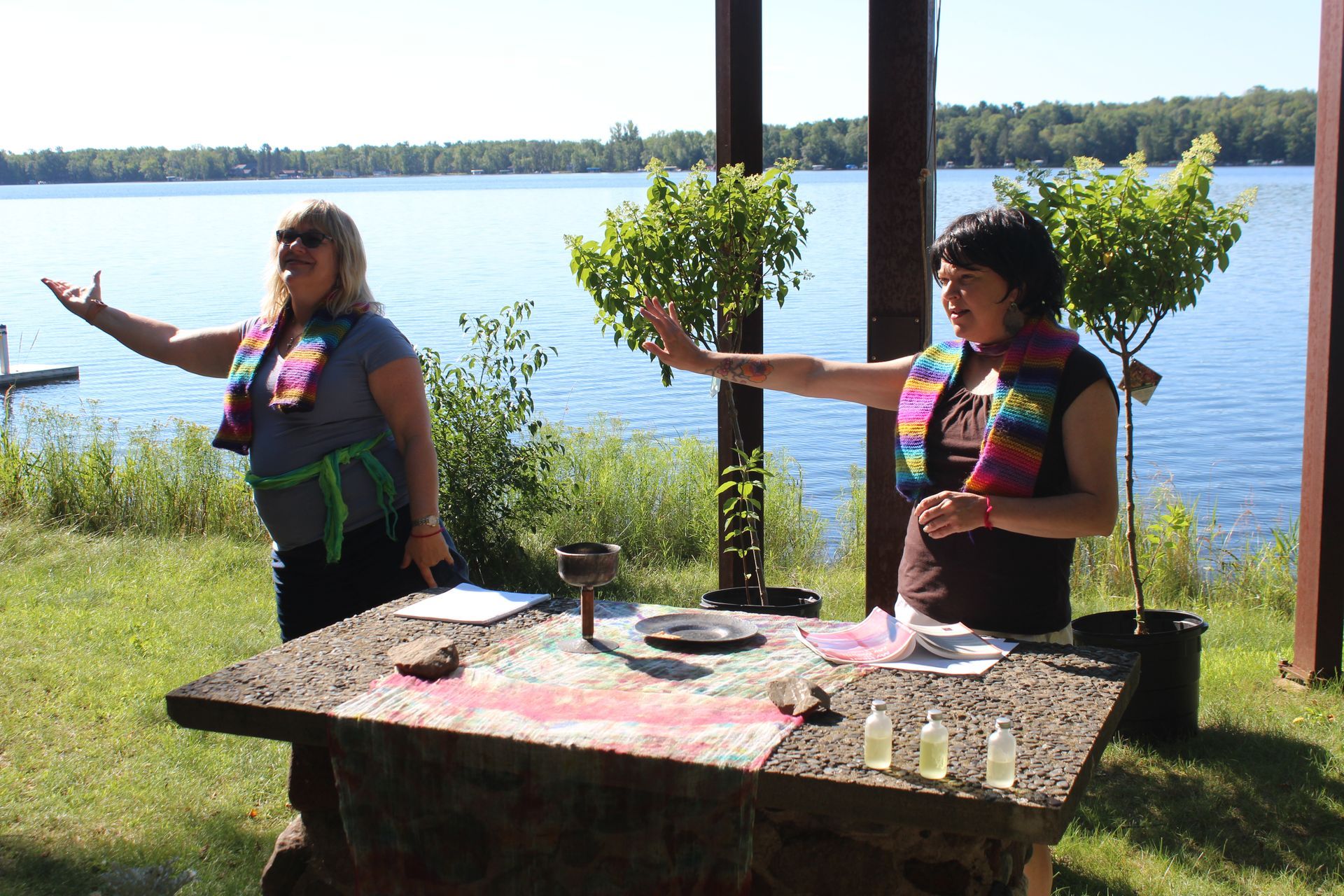 Two people in colorful shirts stand by a lake, gesturing beside a decorated outdoor table.