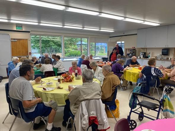 People seated at round tables in a bright community dining hall, with walkers and windows in the background