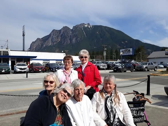 Five people posing outdoors in front of a mountain and parked cars on a sunny day