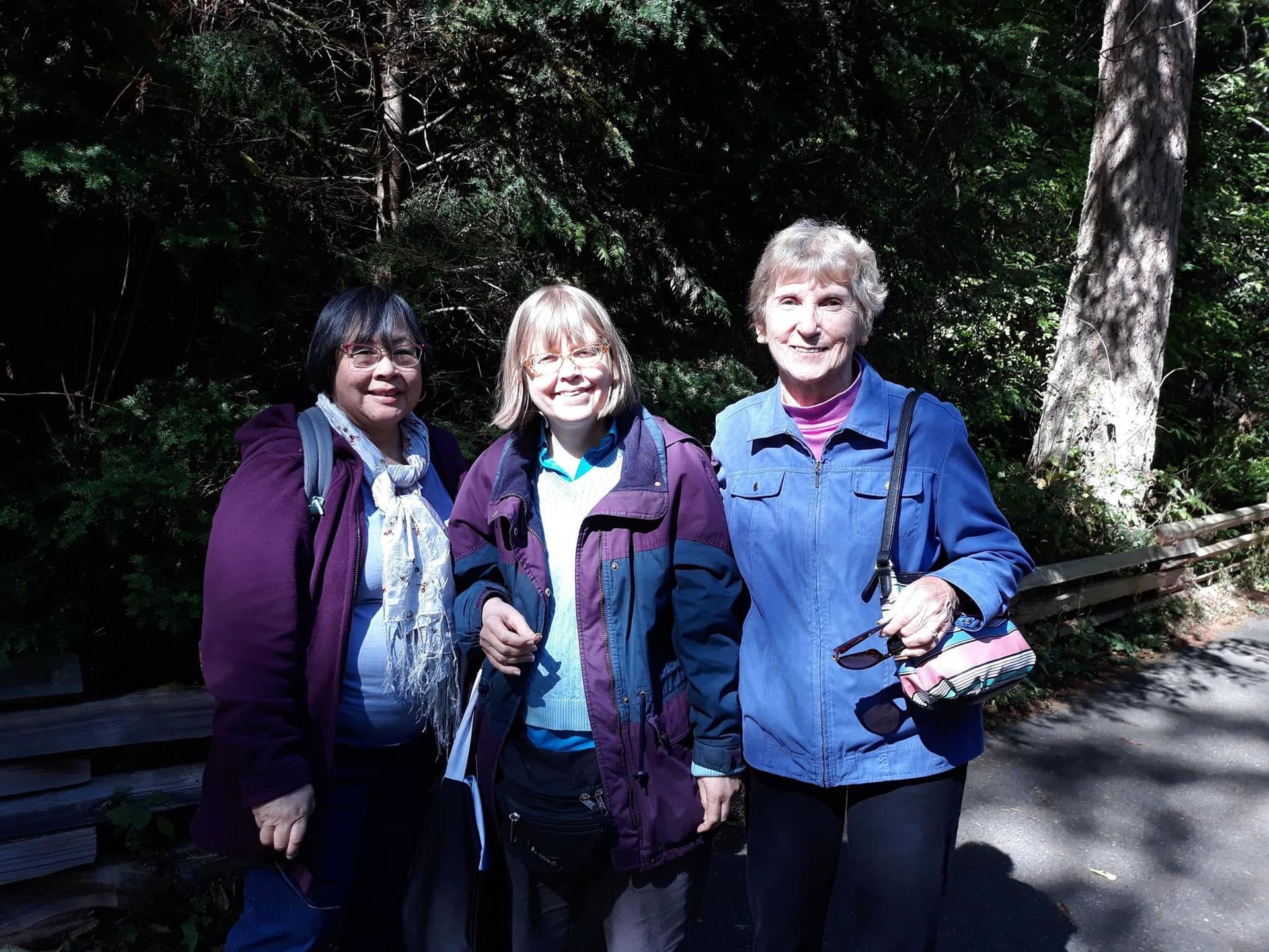 Three smiling people in jackets standing on a sunny wooded trail