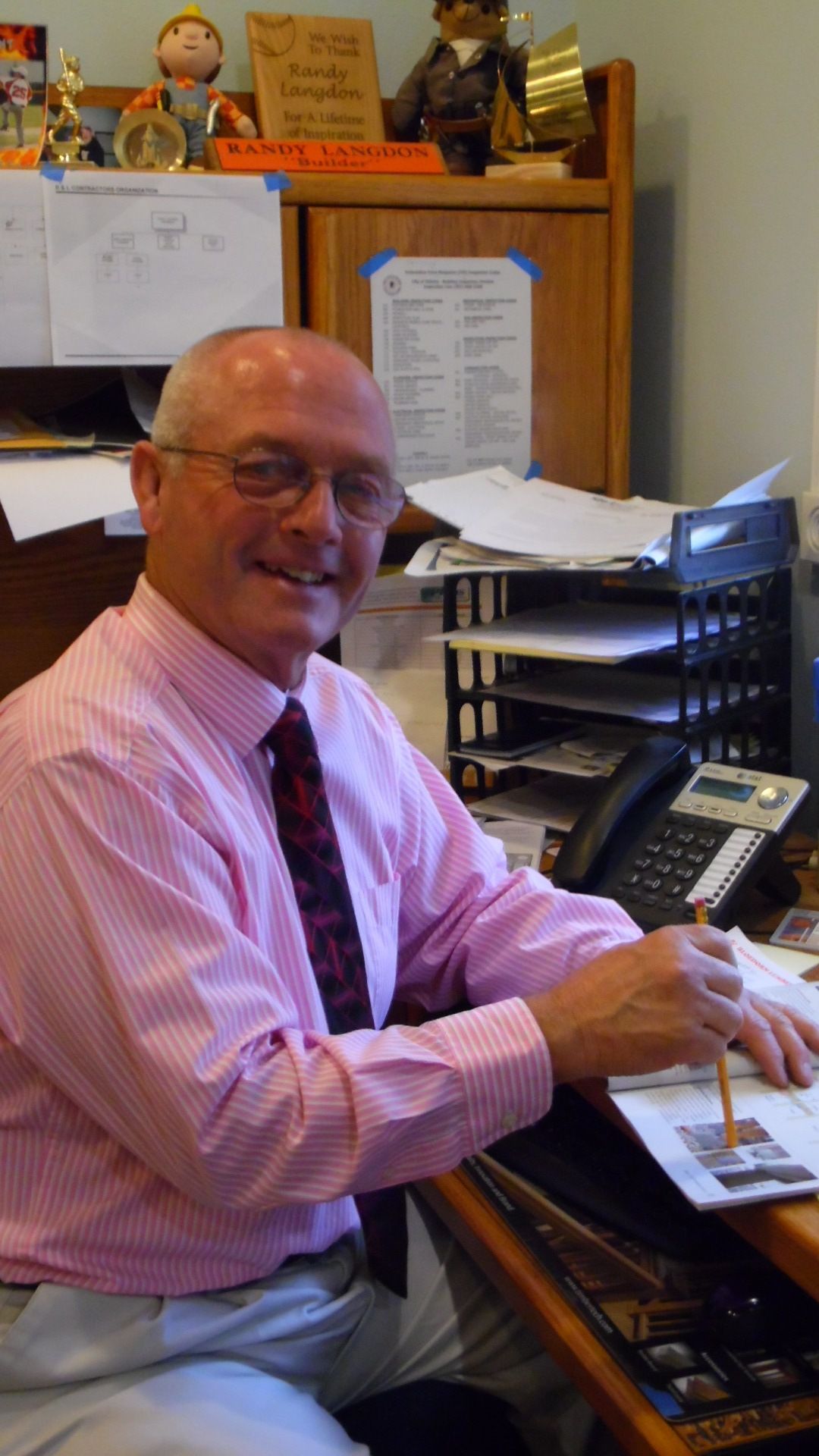 Smiling man in pink shirt and tie sitting at an office desk with papers and a computer monitor