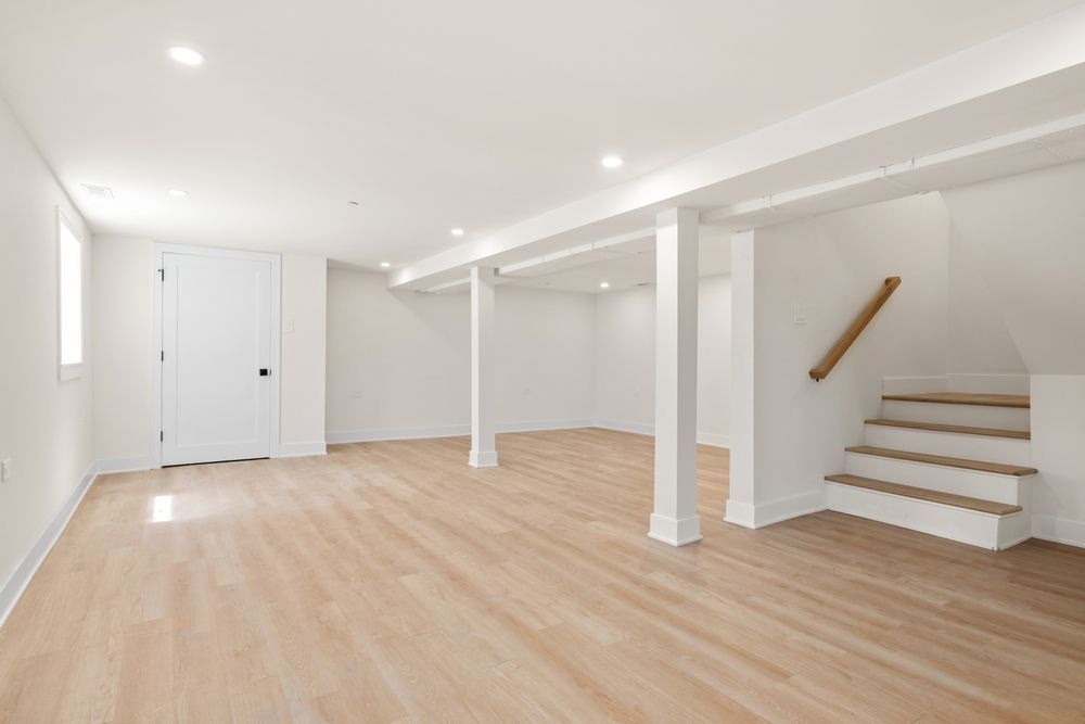 Bright empty basement room with light wood floors, white walls, support columns, and stairs on the right