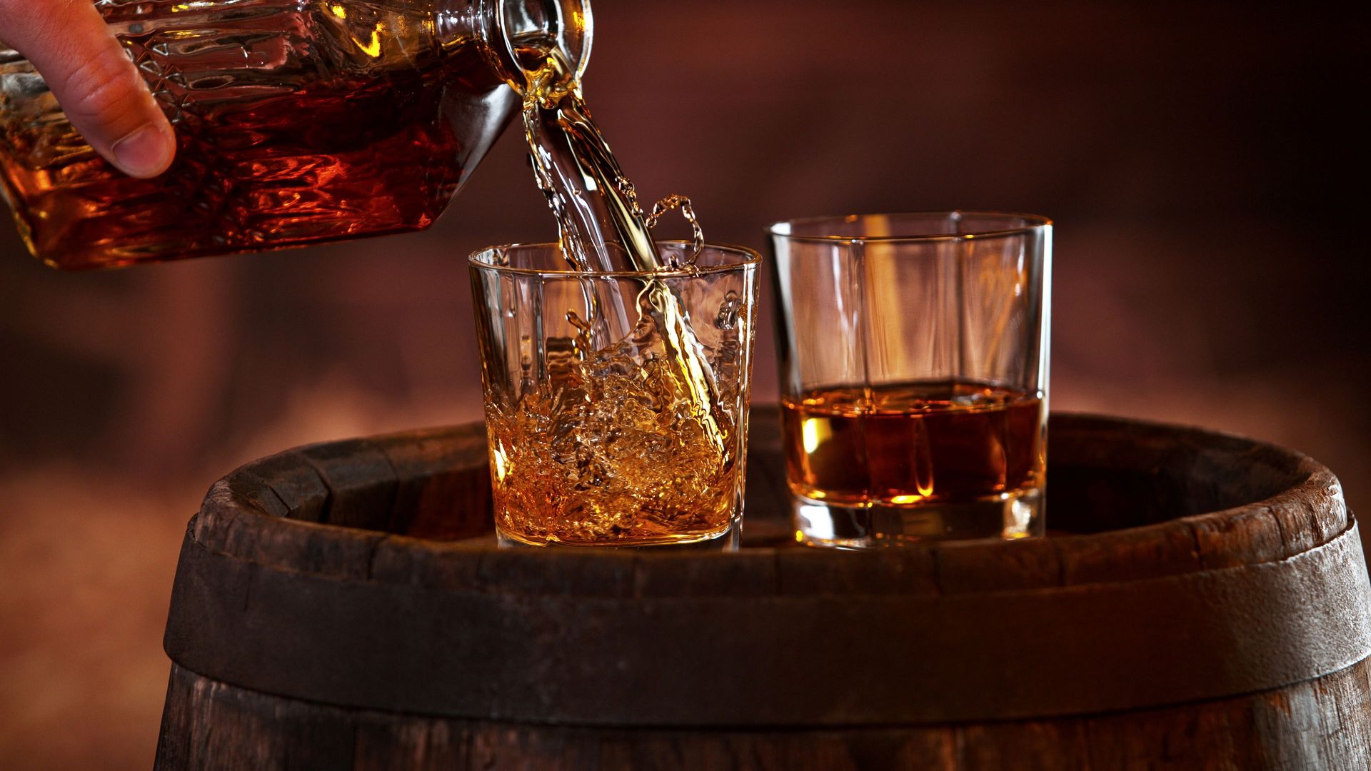 Whiskey being poured from a decanter into two glasses, set on a wooden barrel.