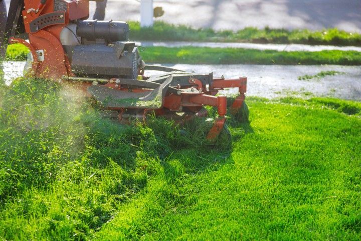 A red commercial lawn mower cuts a strip of tall grass, leaving a neatly mowed path on a sunny day.