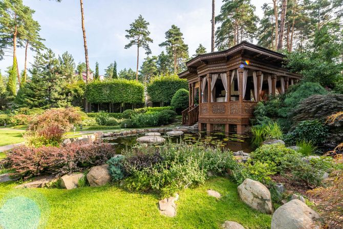 A rustic, wooden gazebo with draped fabric sits beside a small pond in a lush, green landscaped garden.