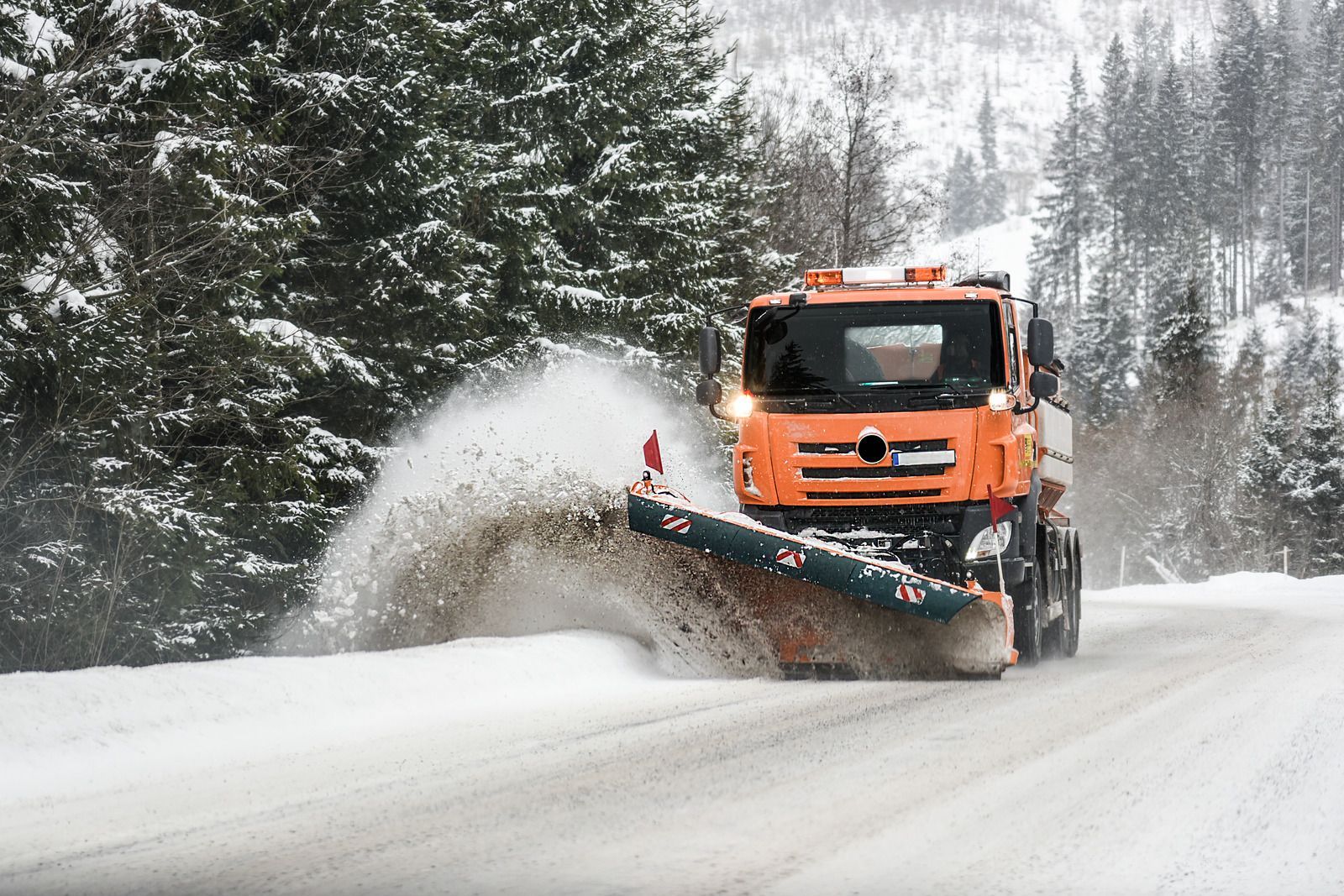An orange snow plow is plowing snow on a snowy road.