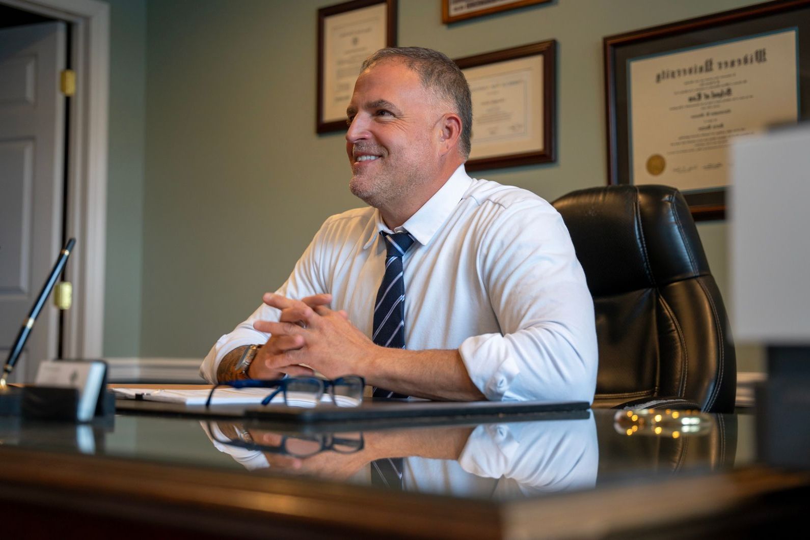 A man in a white shirt and tie is sitting at a desk with his hands folded.