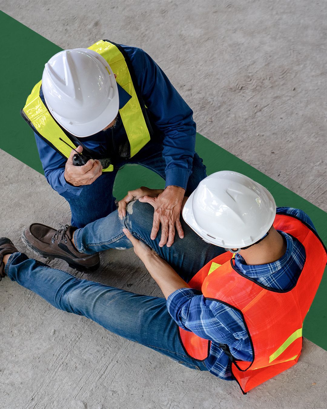 Two construction workers wearing hard hats and safety vests on a concrete floor; one checks the other's injured knee.