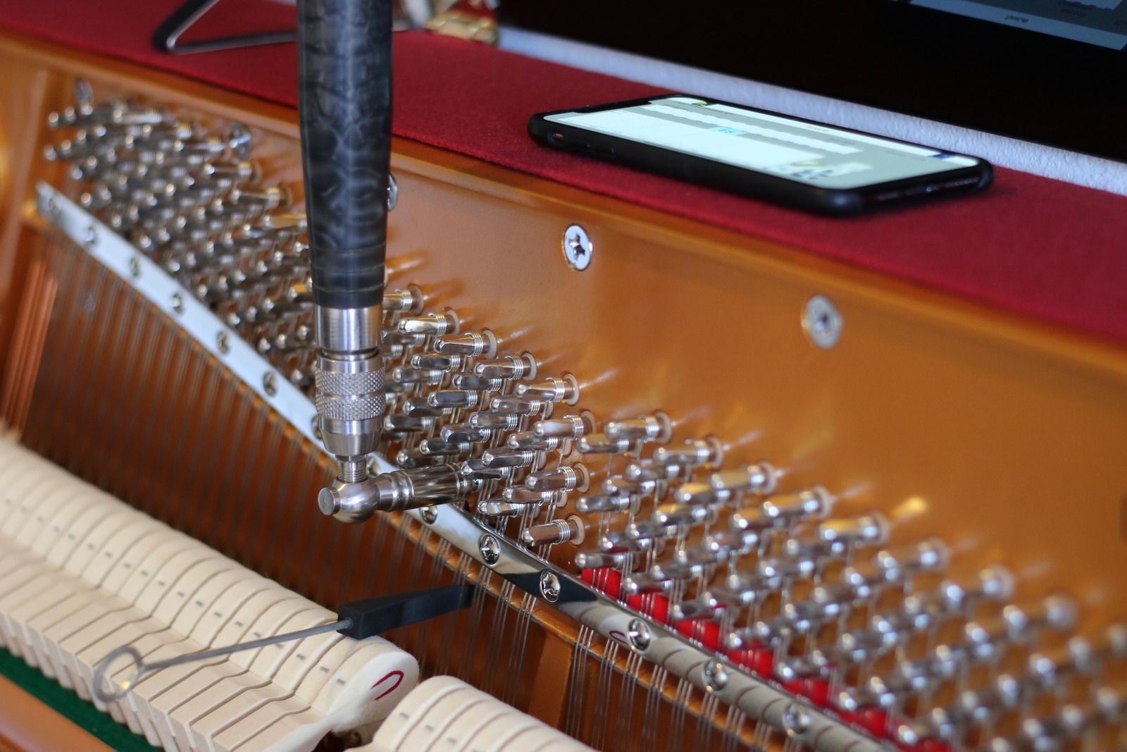 A piano technician uses a tuning tool on piano strings, a smartphone rests nearby.