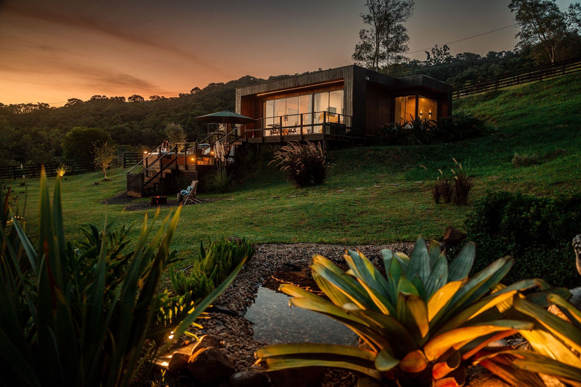 Vista do pôr do sol de uma cabana moderna aninhada em uma encosta verdejante com um pequeno lago.
