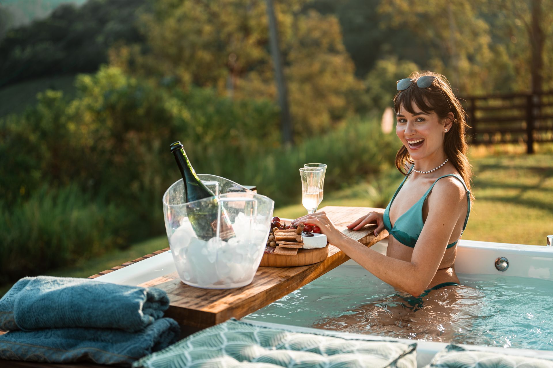 Uma pessoa sorridente em uma piscina segura uma taça de champanhe ao lado de uma bandeja de madeira com um balde de gelo e petiscos ao pôr do sol.