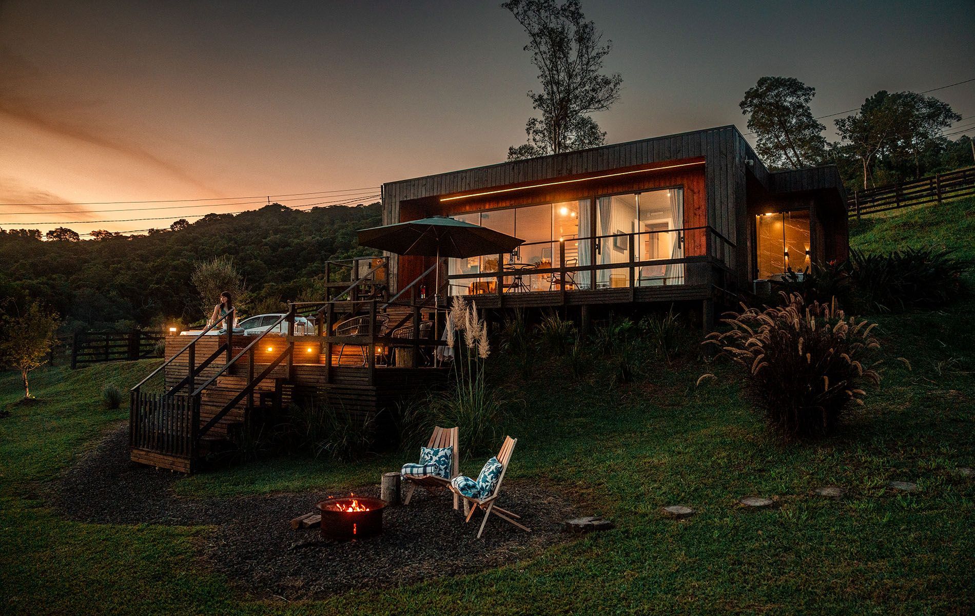 Uma cabana moderna com paredes de vidro fica situada em uma colina gramada ao entardecer, com um deck, uma fogueira e duas cadeiras.