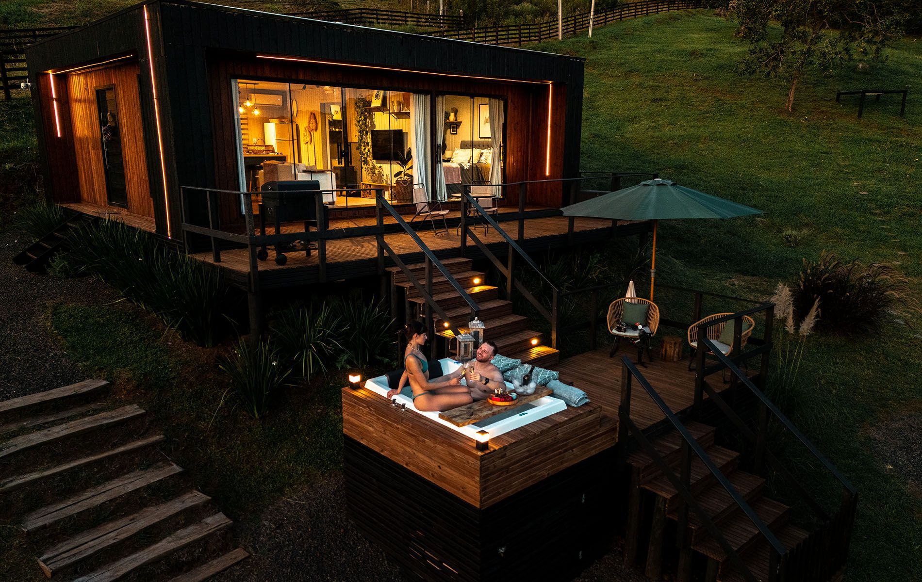 Um casal relaxa em uma banheira de hidromassagem em um deck de madeira ao lado de uma cabana moderna e iluminada, em um ambiente natural e gramado ao entardecer.