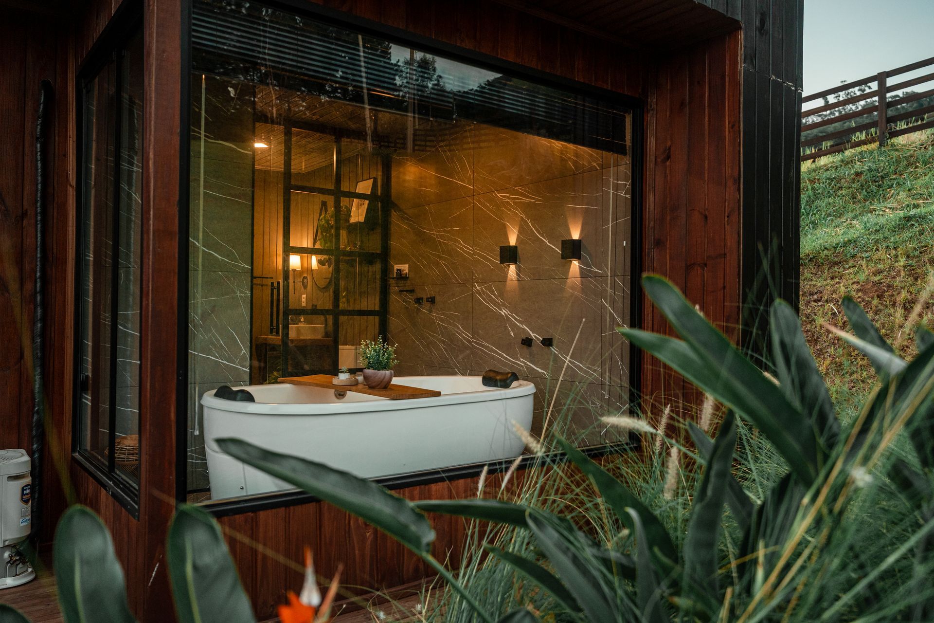 Uma banheira branca fica dentro de uma cabana de madeira com grandes janelas de vidro, com vista para uma colina gramada ao entardecer.