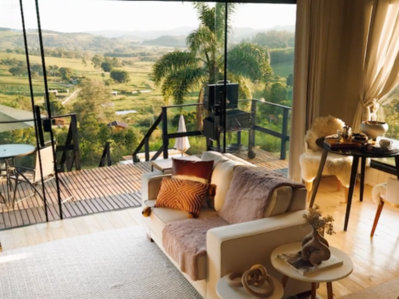 Sala de estar com sofá, mesa e janelas do chão ao teto com vista para uma paisagem montanhosa e panorâmica e um deck externo.