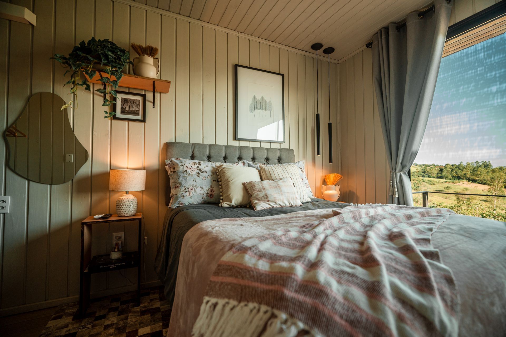 Um quarto aconchegante com cama, paredes verticais de madeira em tons claros, luminárias pendentes, uma pequena mesa de cabeceira e uma grande janela.