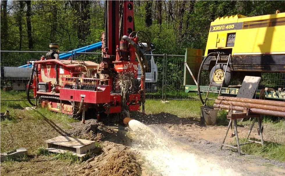 Una piattaforma di perforazione rossa con spruzzi d'acqua, in un'area erbosa, perfora il terreno. Sullo sfondo, un generatore giallo.
