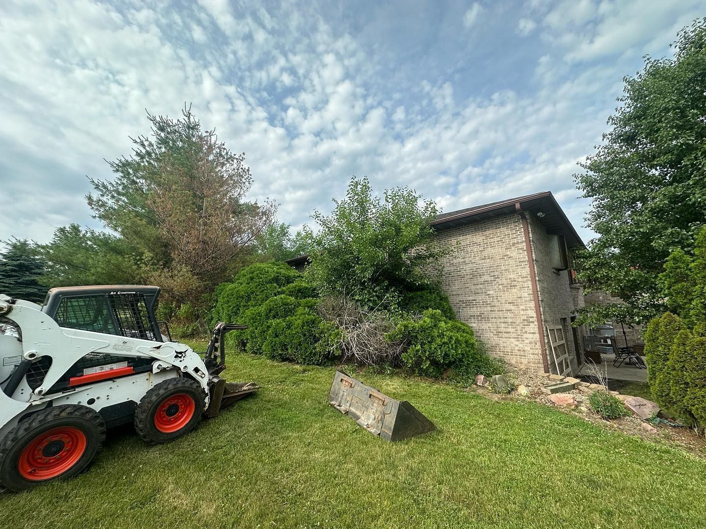 A white Bobcat clearing overgrown greenery near a stone building on a sunny day.