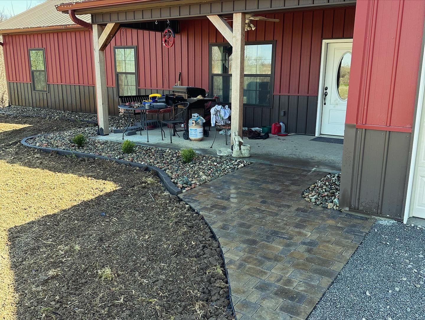 Brick pathway leading to a red and brown house with a porch. Landscaping, grass, and gravel border the pathway.