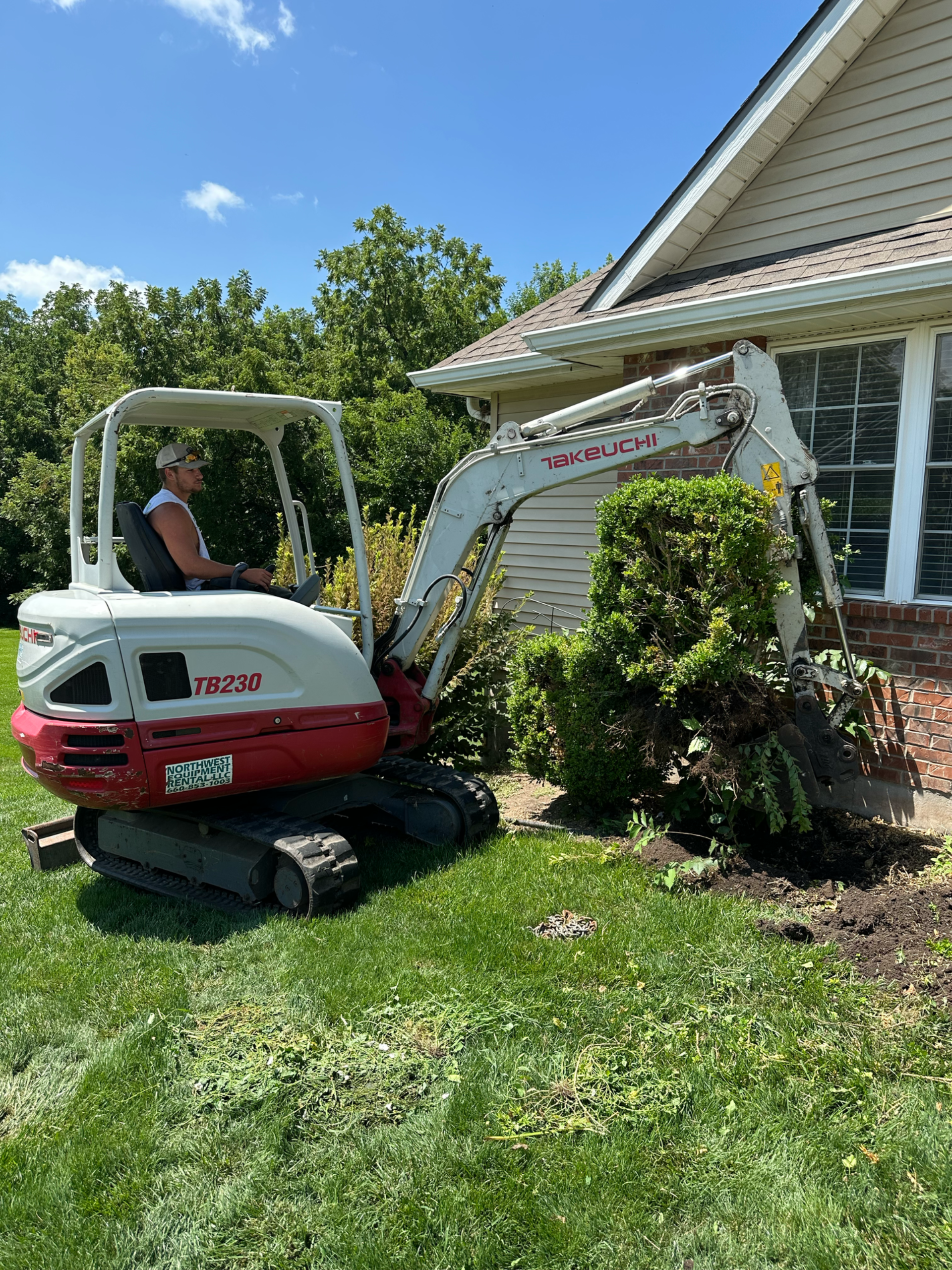 Man operates a small excavator, removing bushes next to a house. Bright sunlight, green grass.