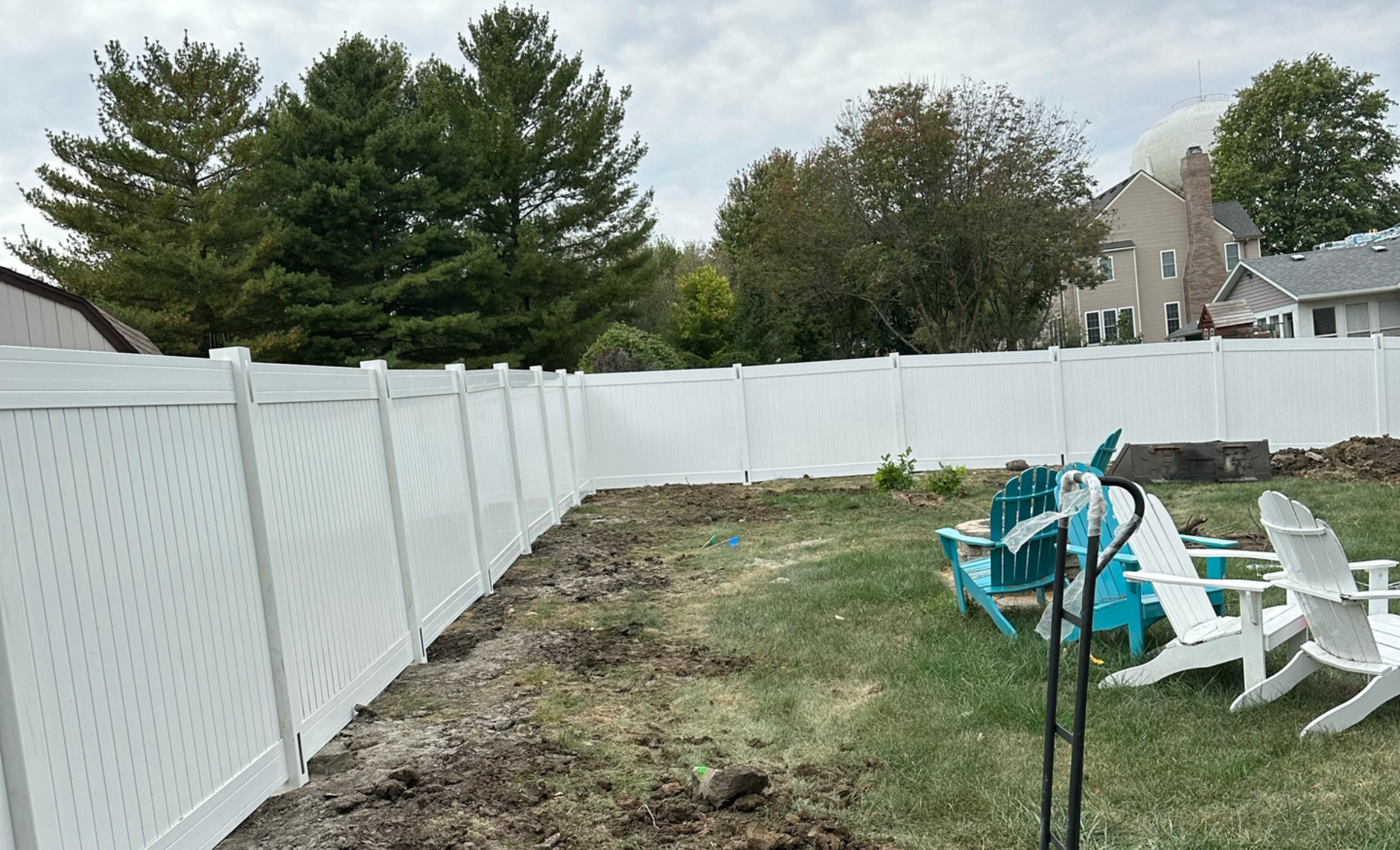 White vinyl fence encloses a yard with dirt, grass, and Adirondack chairs. Trees and a house are in the background.