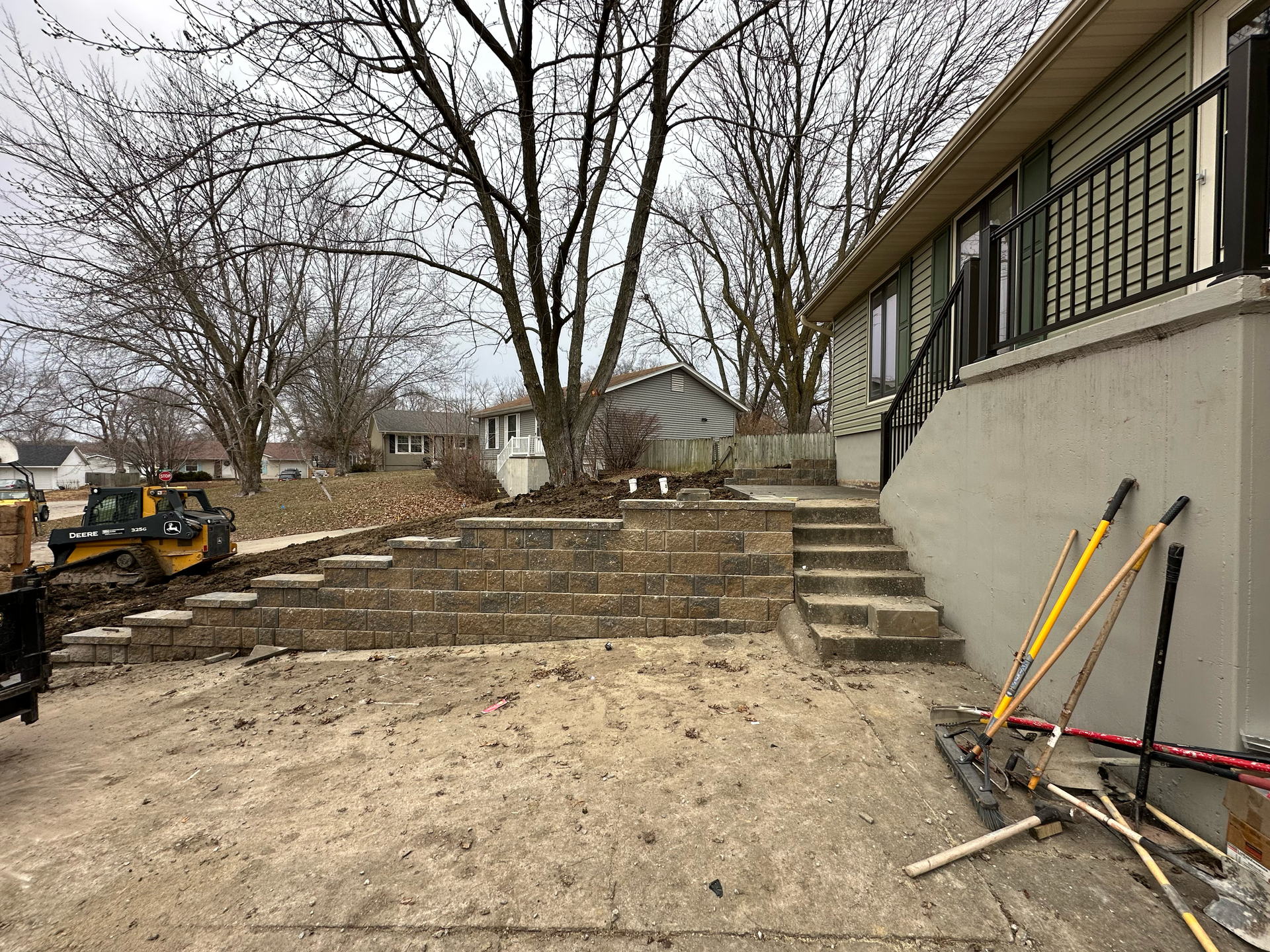 Construction site with tiered retaining wall and steps next to a house; tools are leaning on the wall.