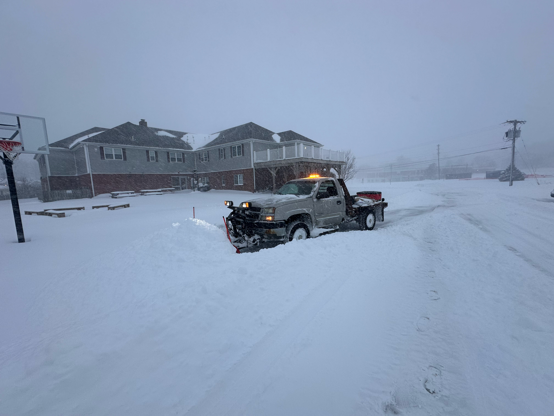 Snow plow clearing a snowy road in front of a house during a blizzard.