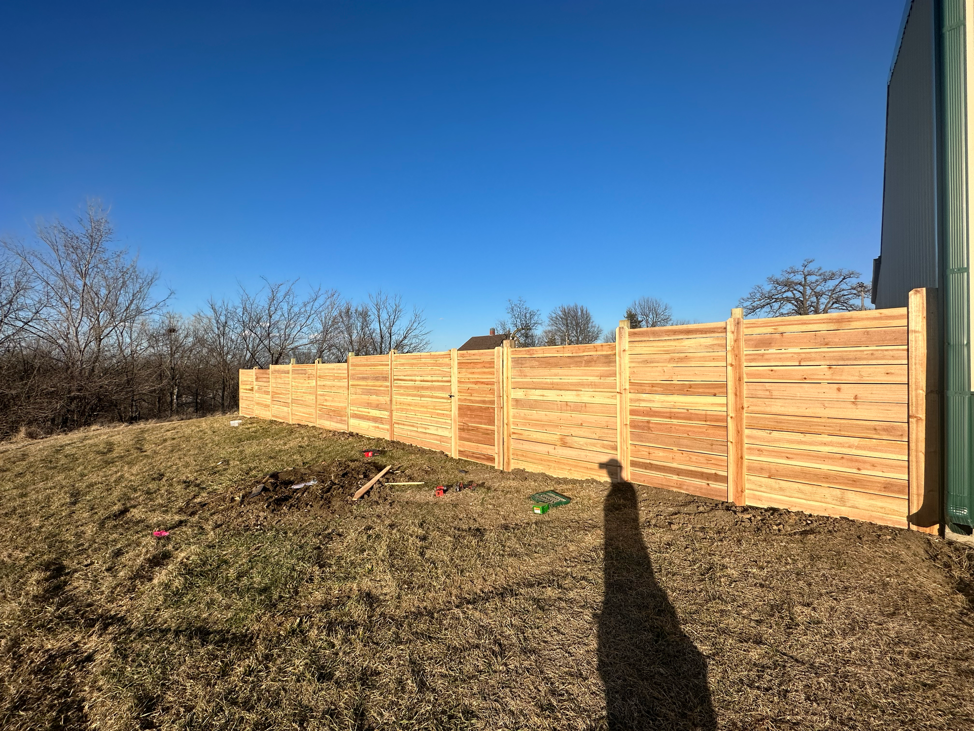 Wooden fence in a grassy field under a clear blue sky. Shadow of person visible.