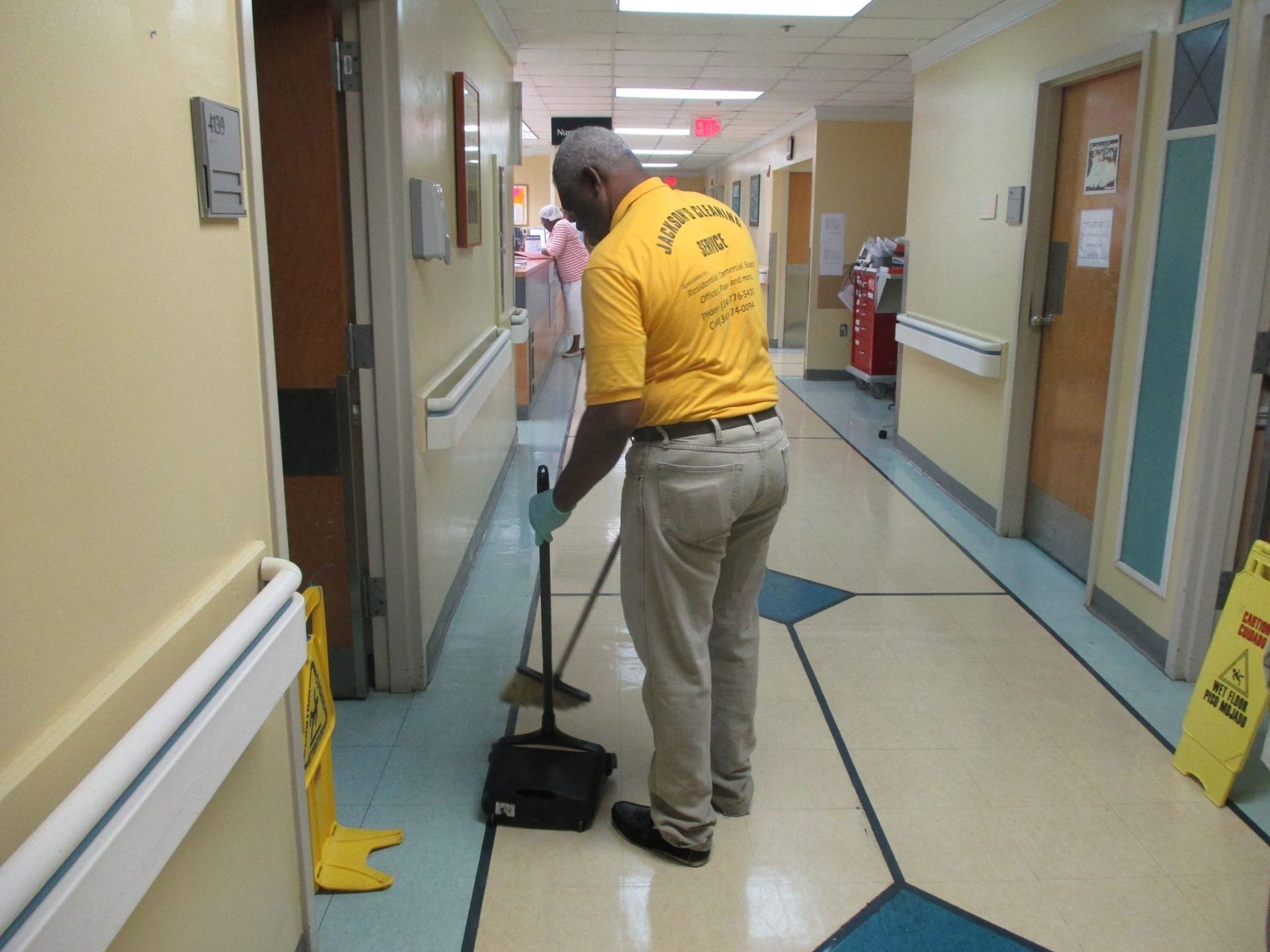 Person sweeping hospital hallway with dustpan and broom. Yellow caution signs and doors visible.