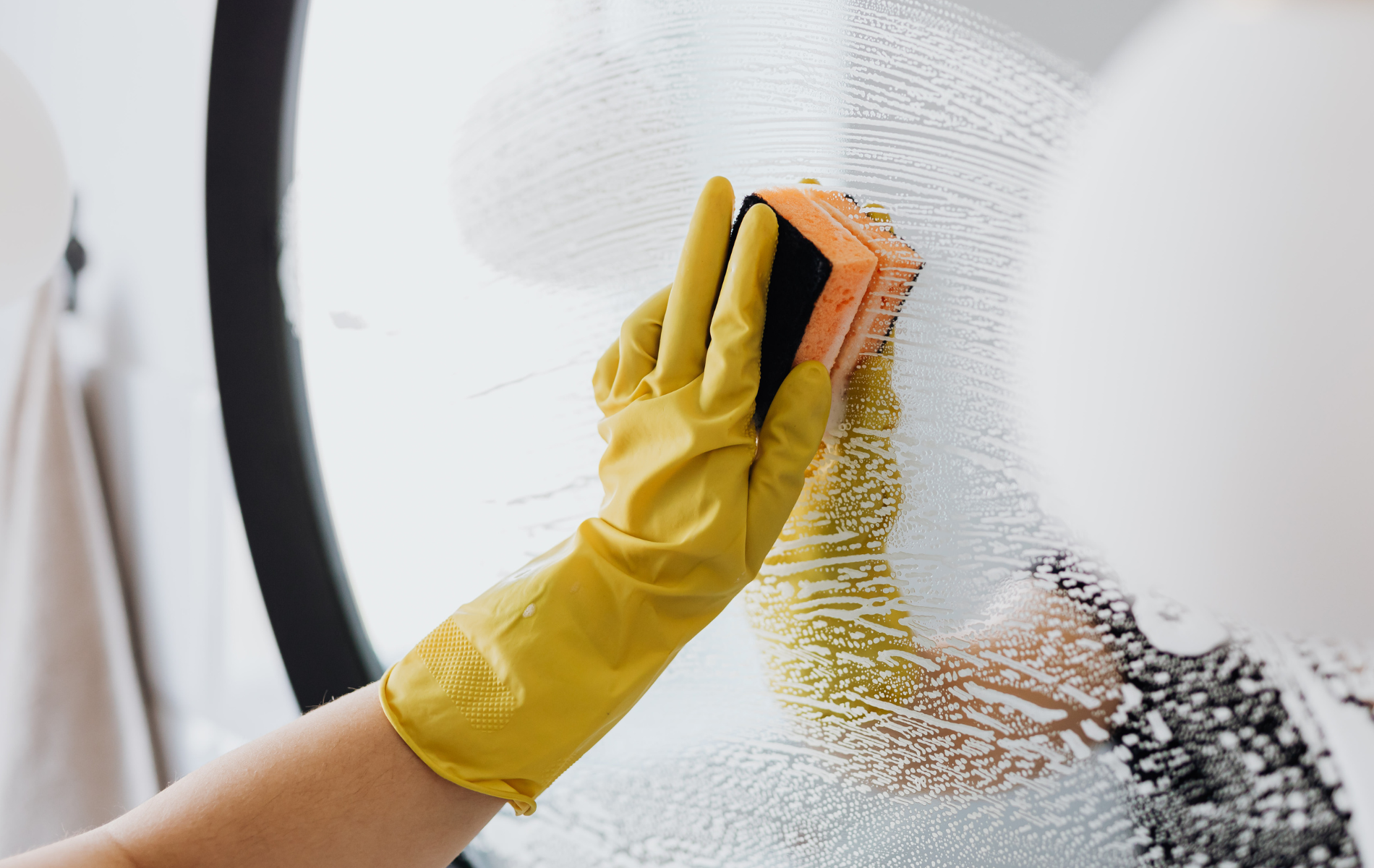 Person wearing yellow gloves cleaning a mirror with a sponge.
