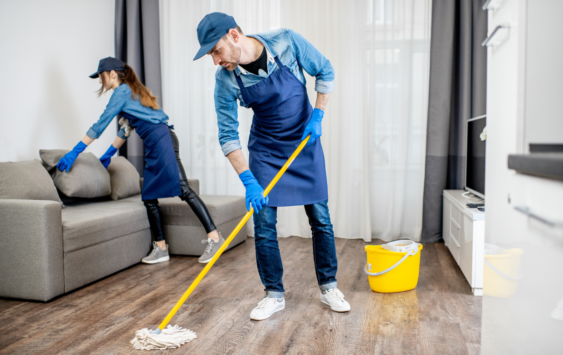 Two people cleaning a room: one mopping, the other arranging a couch pillow.