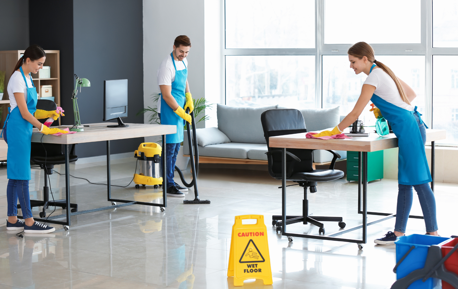 Three cleaners mopping a bright office floor around desks and a wet floor sign
