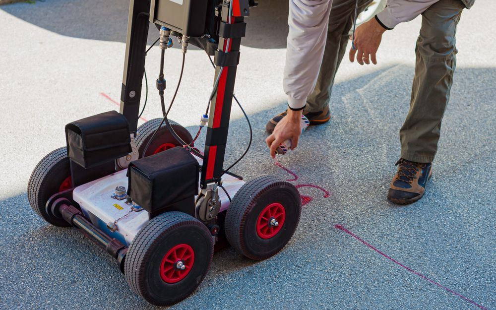 Person uses spray paint to mark a line, next to a wheeled GPR (ground-penetrating radar) device on pavement.
