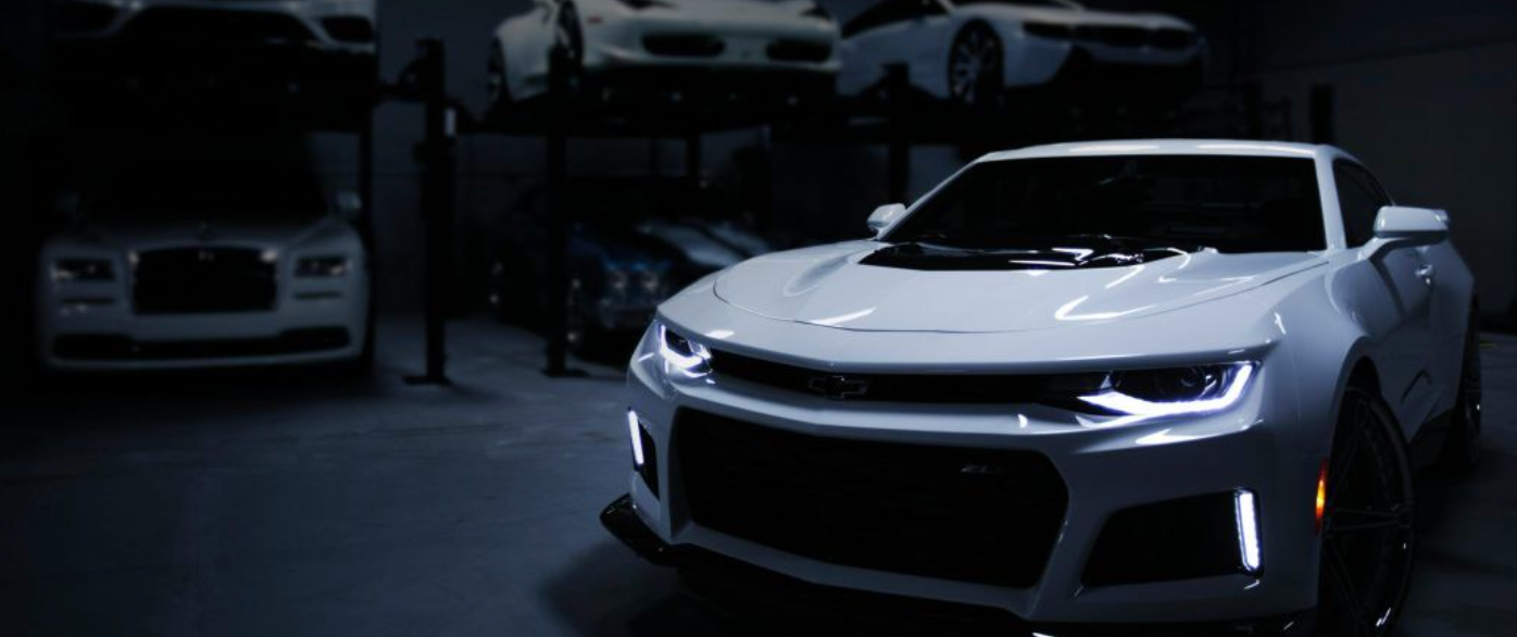 White Chevrolet Camaro in a garage, other cars in the background, dimly lit.